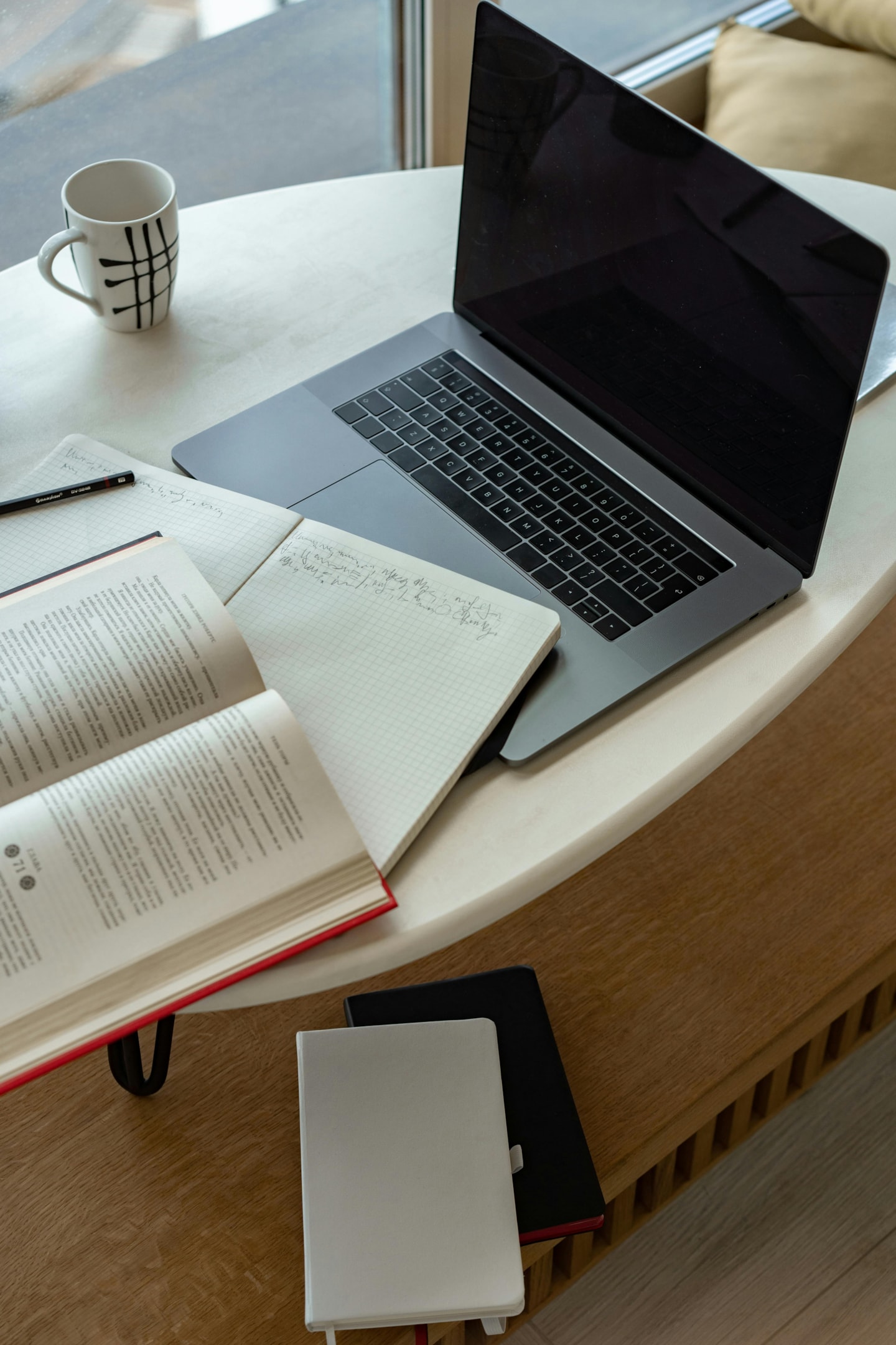 A MacBook pro on a white table, together with a notebook and a textbook open next to it.