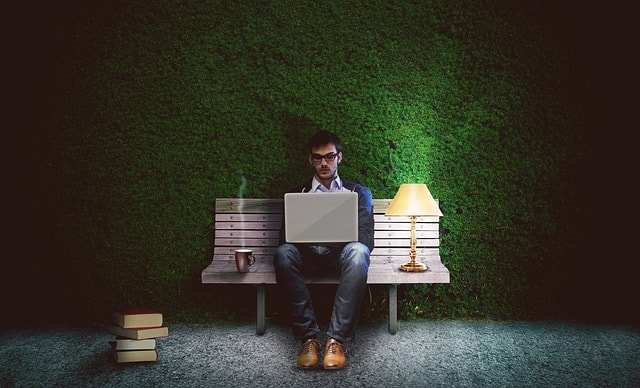 A man sitting between two chairs that have a cup of coffee and a lamp on them and writing on his laptop.