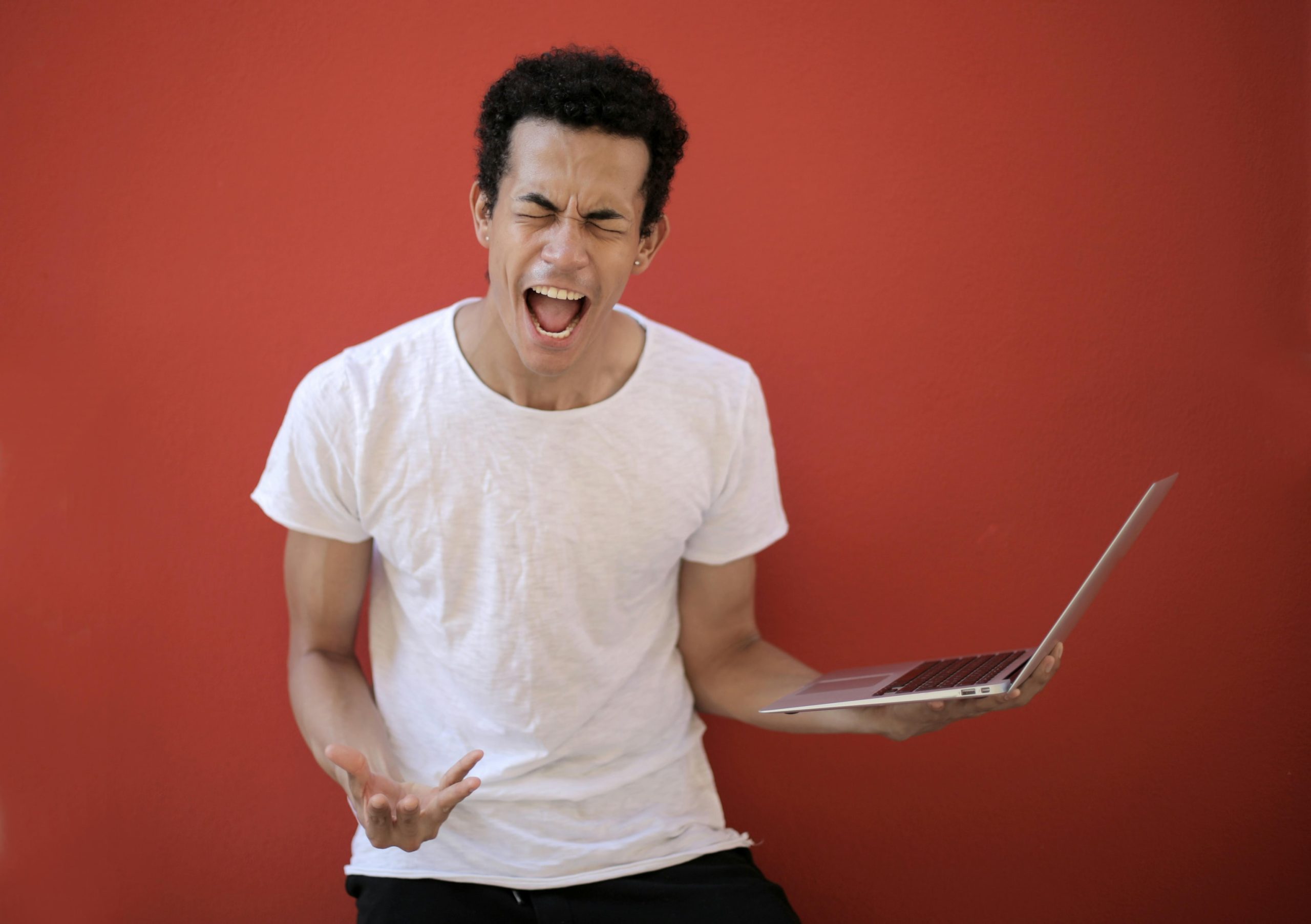 A young man holding a laptop in one hand and making a facial expression to suggest he is shouting. 