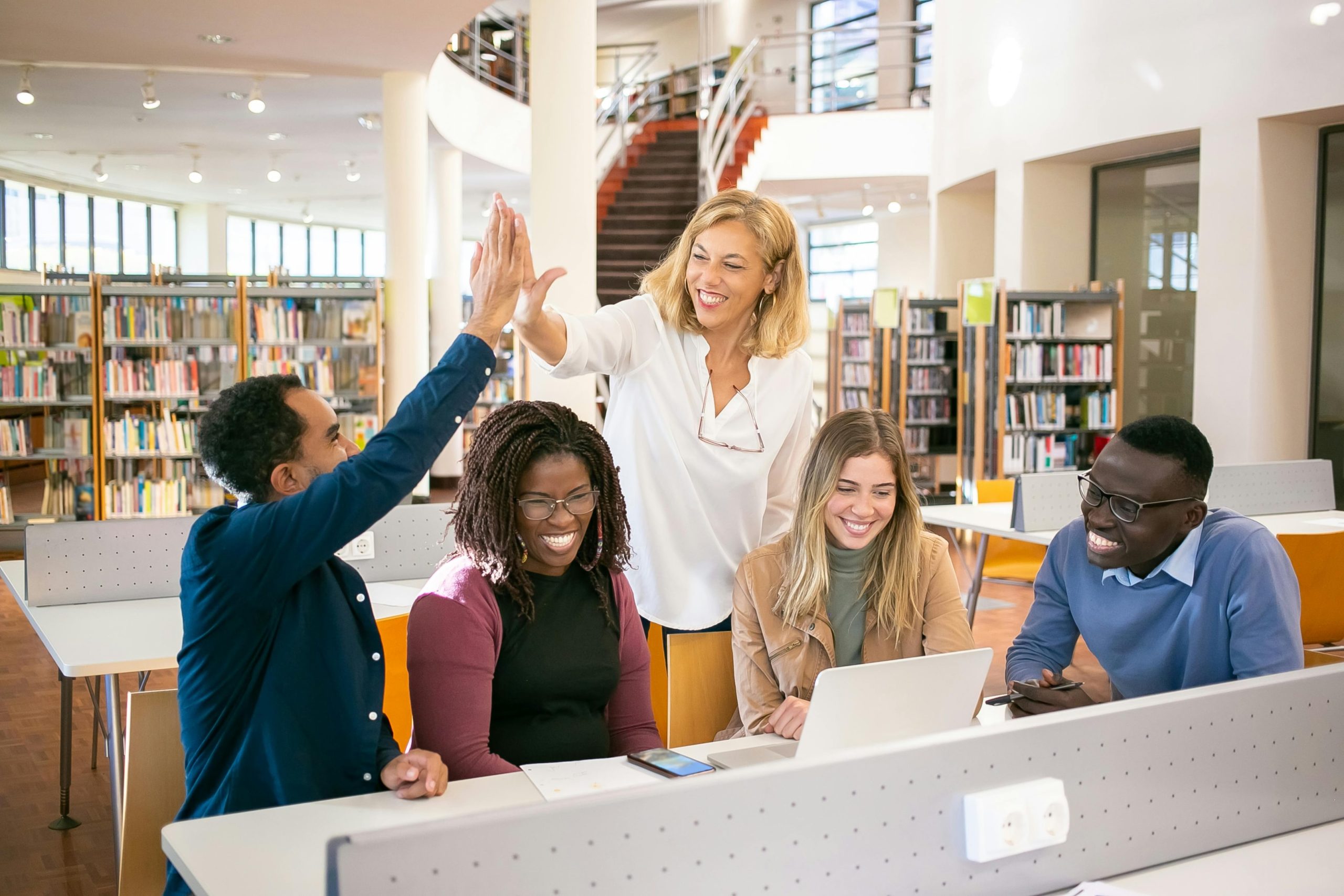 Cheerful students sitting in a library around one laptop. A student and a teacher are high-fiving each other.