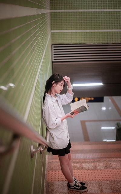 A female student standing on the stairs of her school and reading a book.