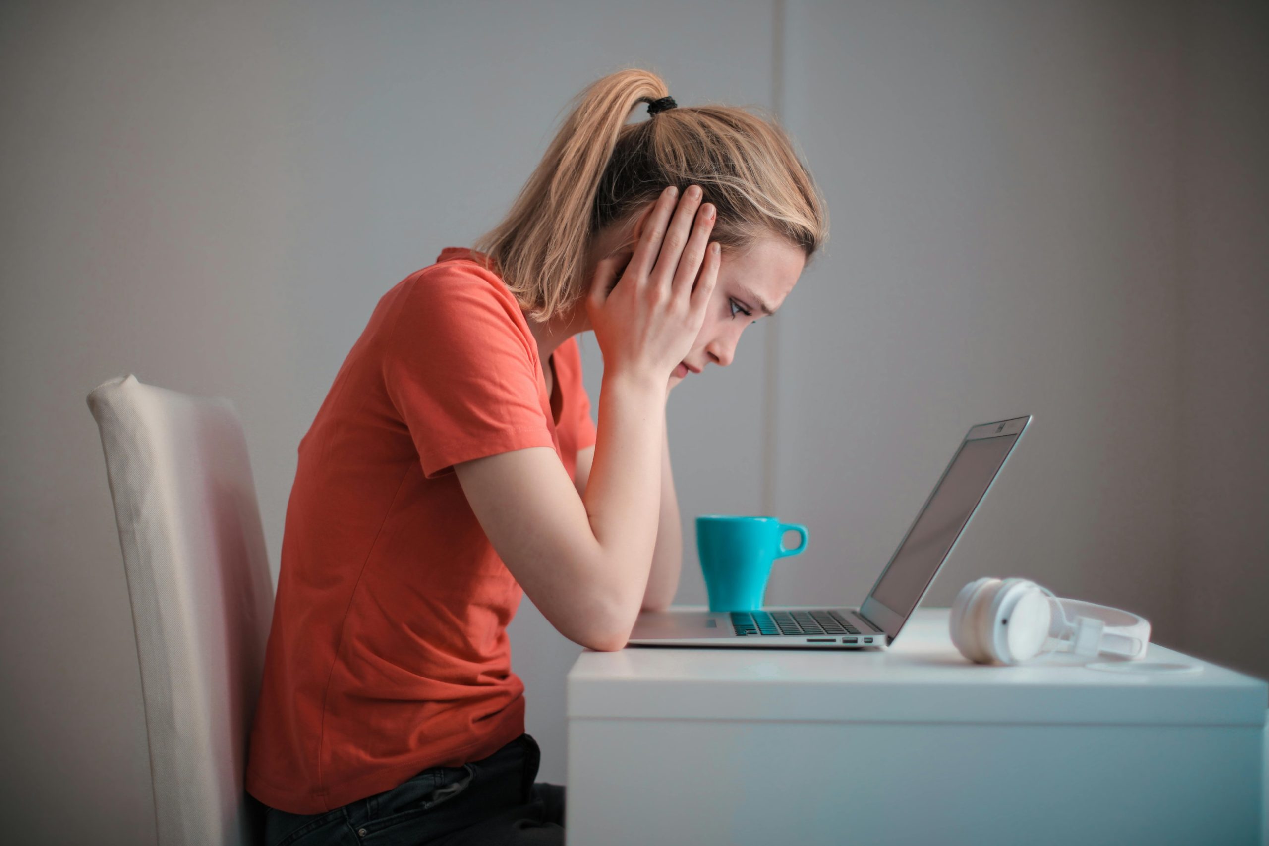 A young woman holds her head in her hands whilst she is sitting in front of her laptop.