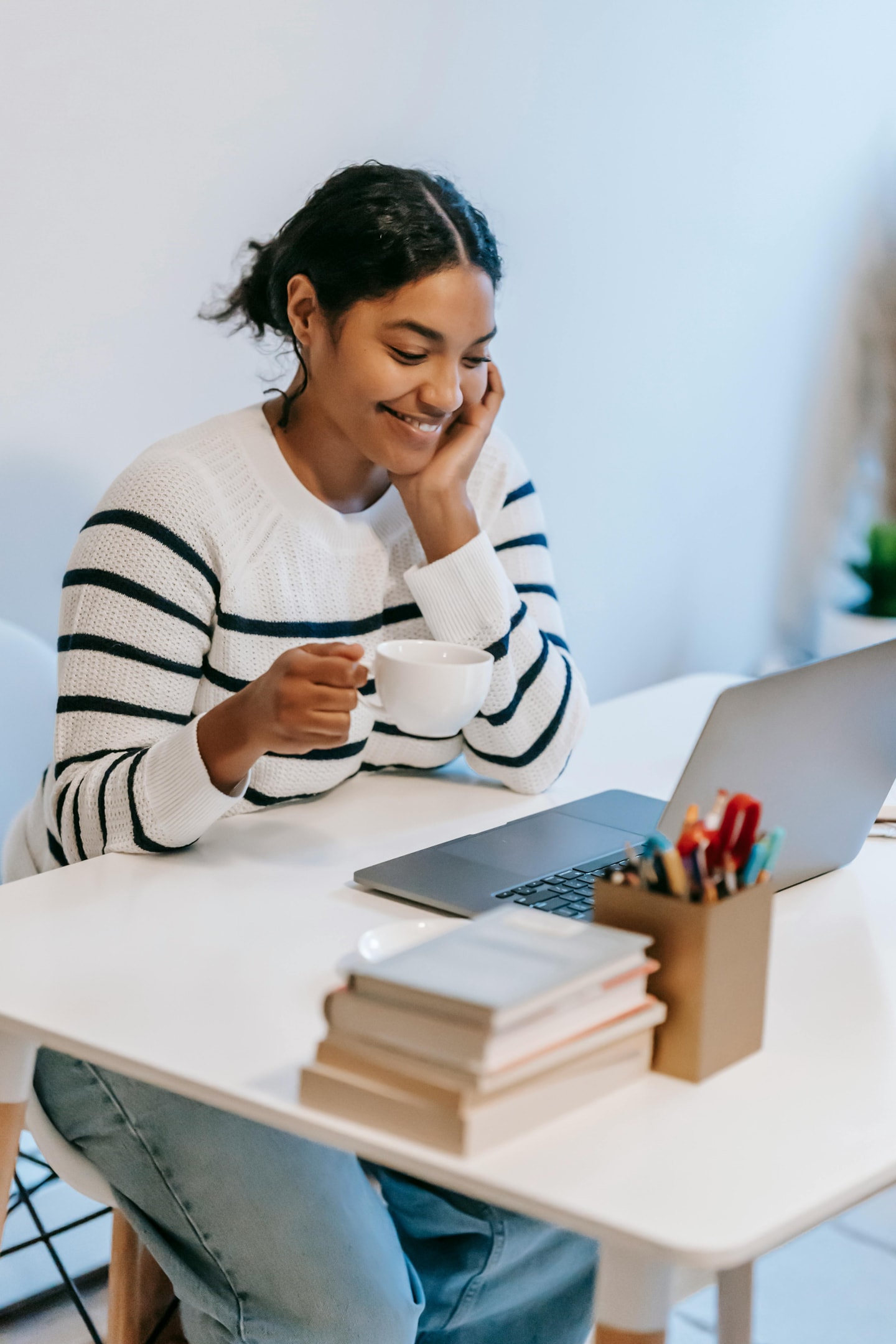 A smiling young woman with a cup of coffee in one hand working on her laptop at a desk.