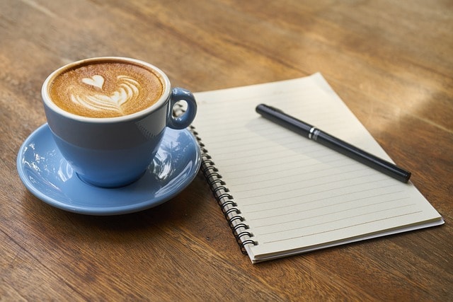 A blue cup of coffee next to a notebook and a black pen.