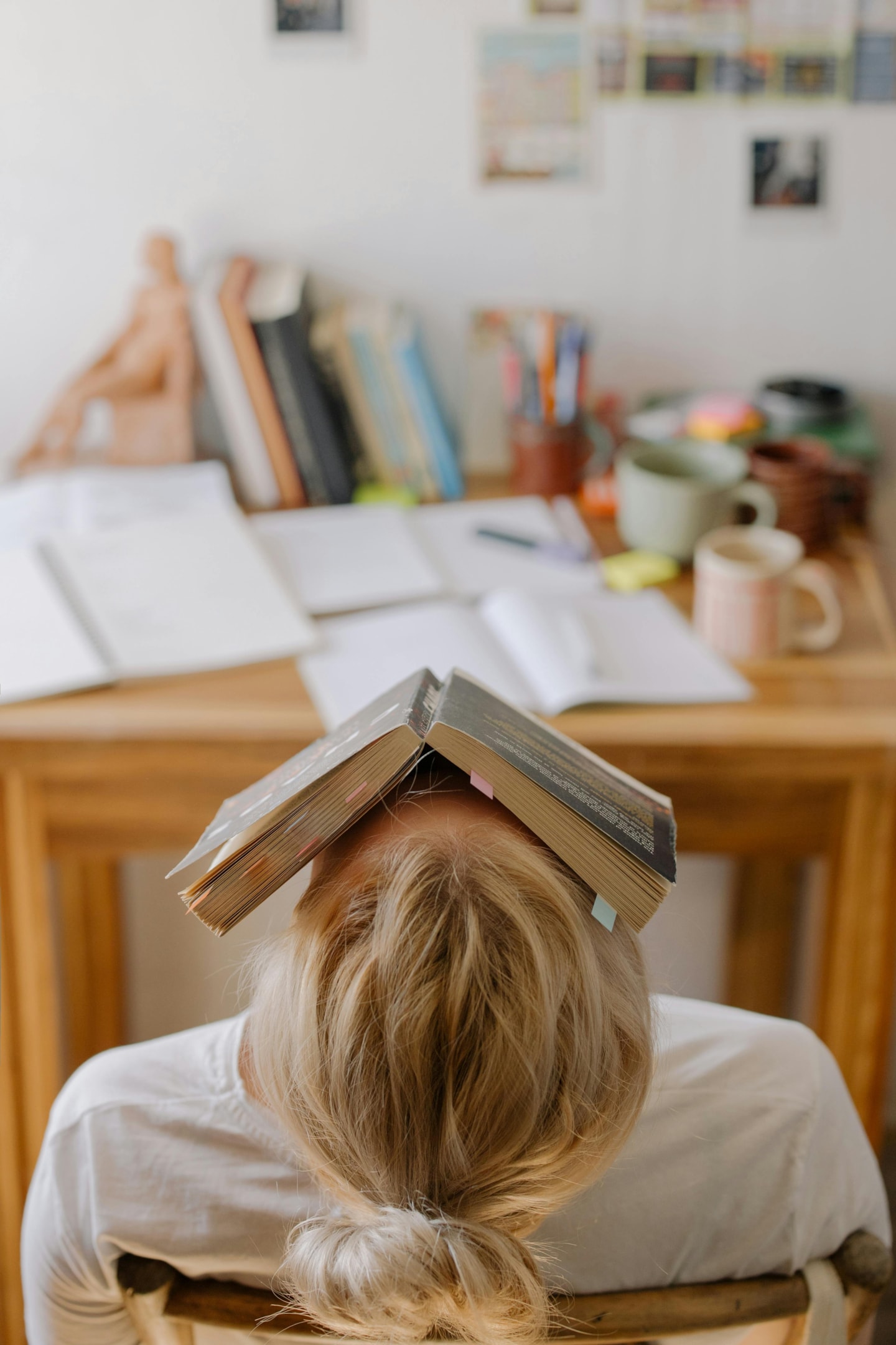 A girl sitting at a desk at home and leaning back while having an open book on her face.