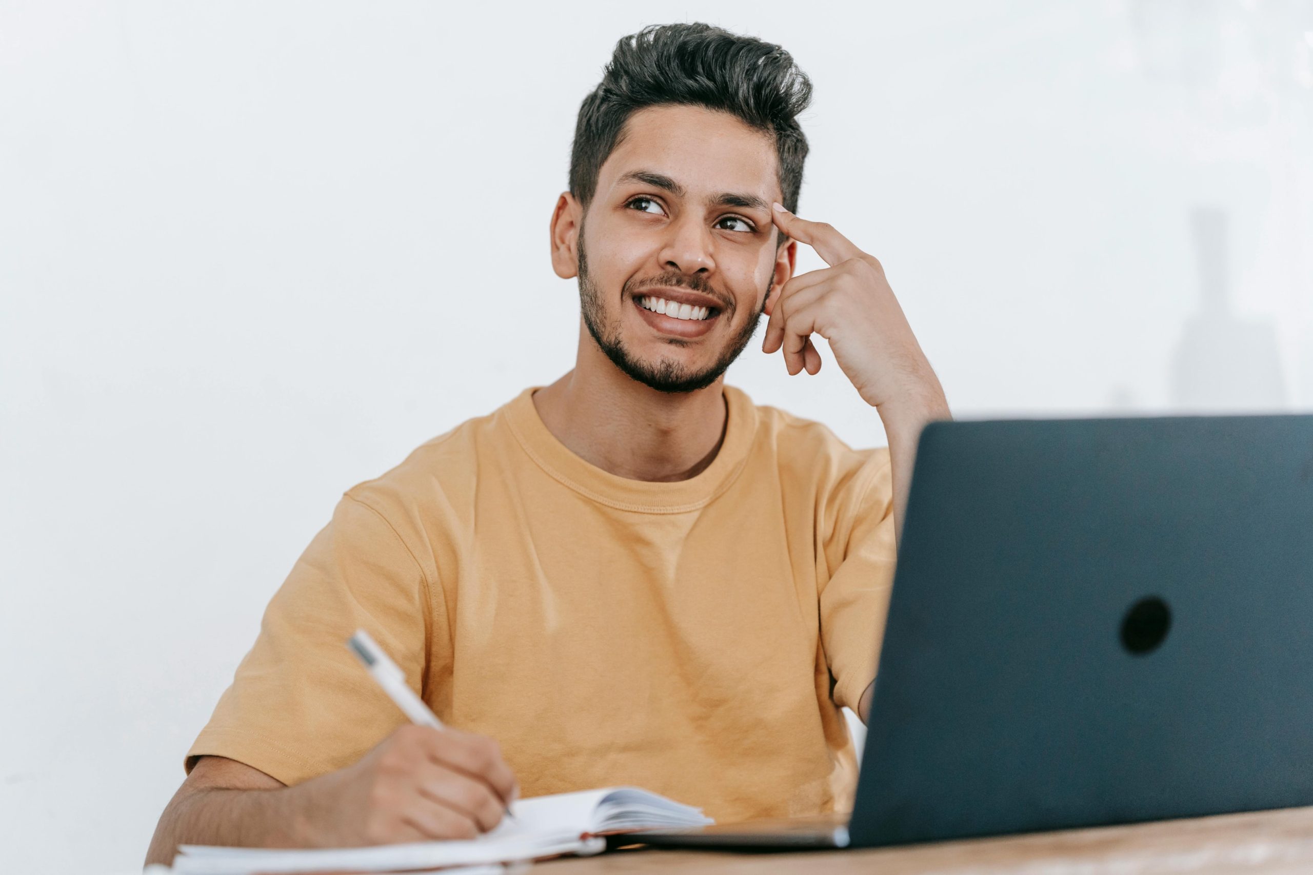 A smiling man working on a laptop and taking notes. 