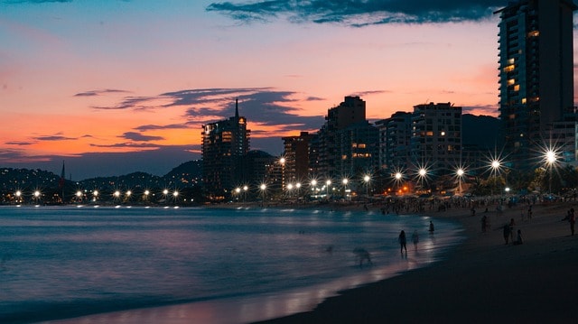 A beach at sunset with buildings with lights in the background.