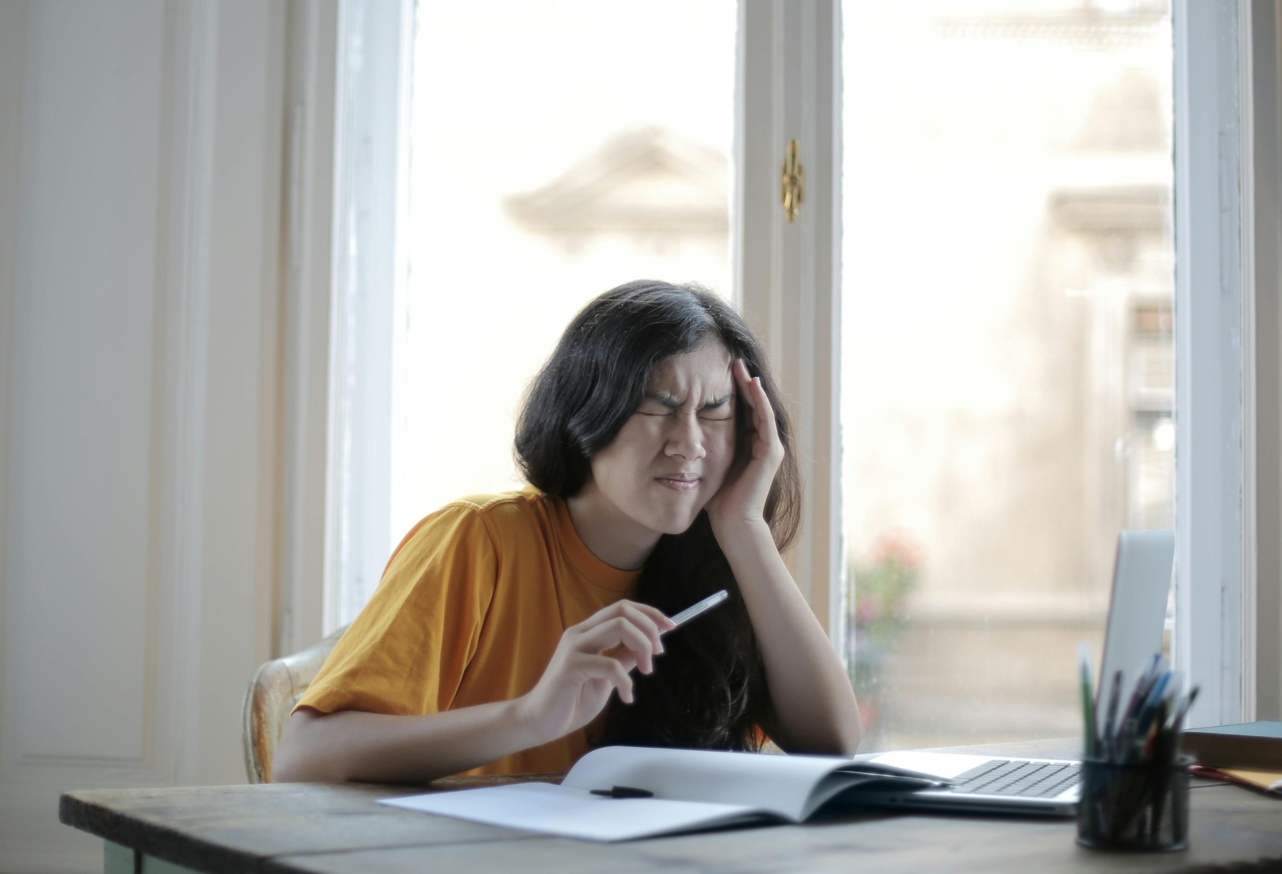 A student holding her head and squinting while sitting at her desk with an open notebook and a pen in her hand.