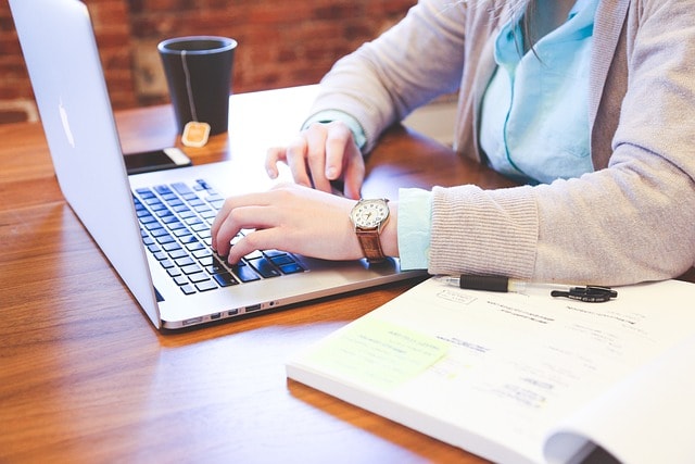 A person typing on a laptop with a cup of tea and a notebook next to them.