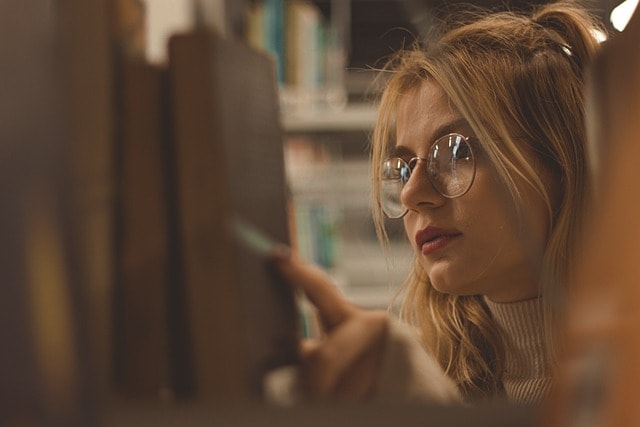 A girl with reading glasses picking a book up from a library shelf.