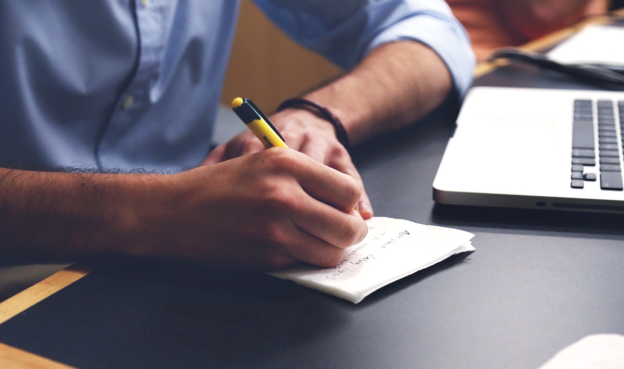 A man sitting on a desk with his laptop and taking notes.