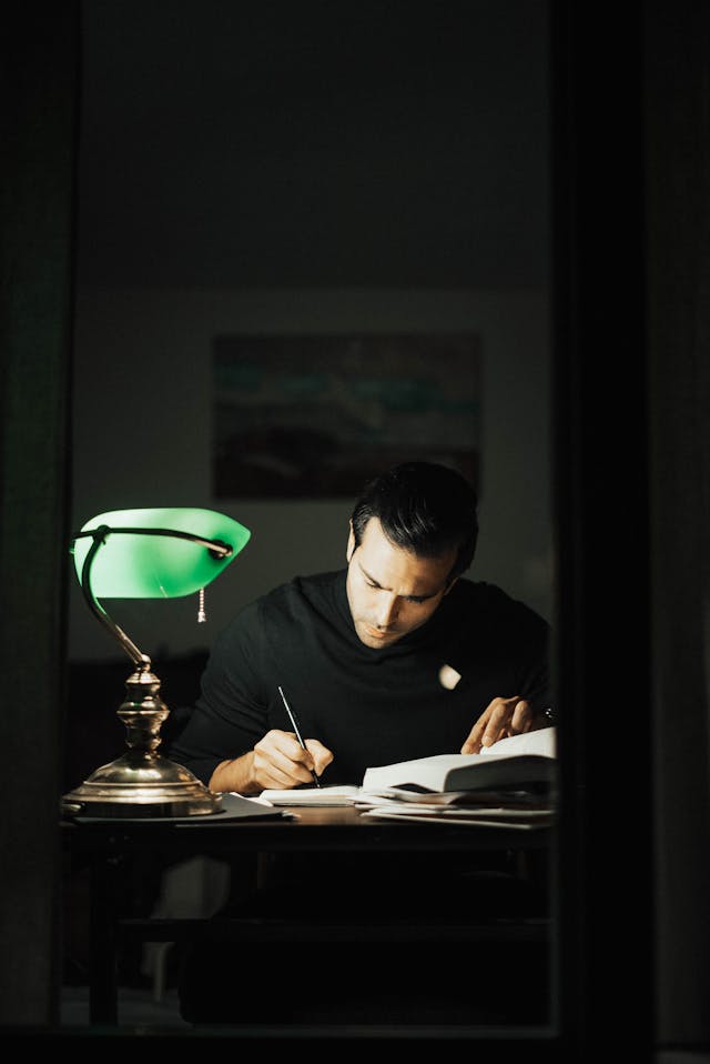 A man at a desk at night with a lamp illuminating his face while he writes in a notebook. 