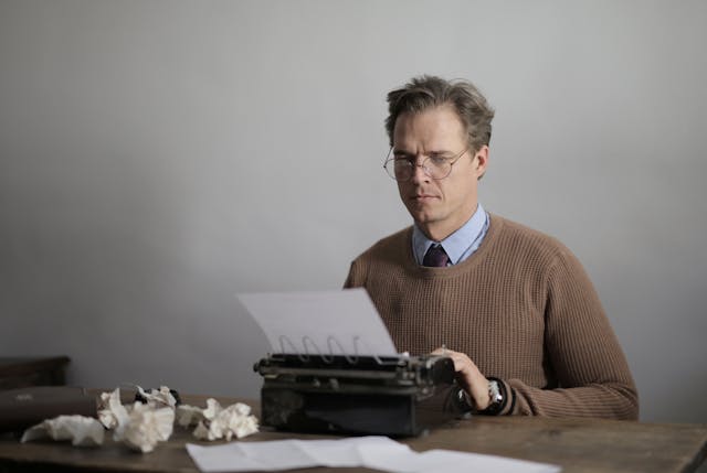 A man in a brown sweater with glasses uses a black typewriter machine. 