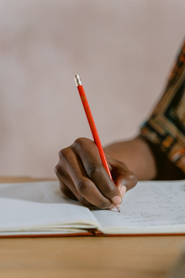 A close-up of a person who holds a red pencil and writes in a notebook.