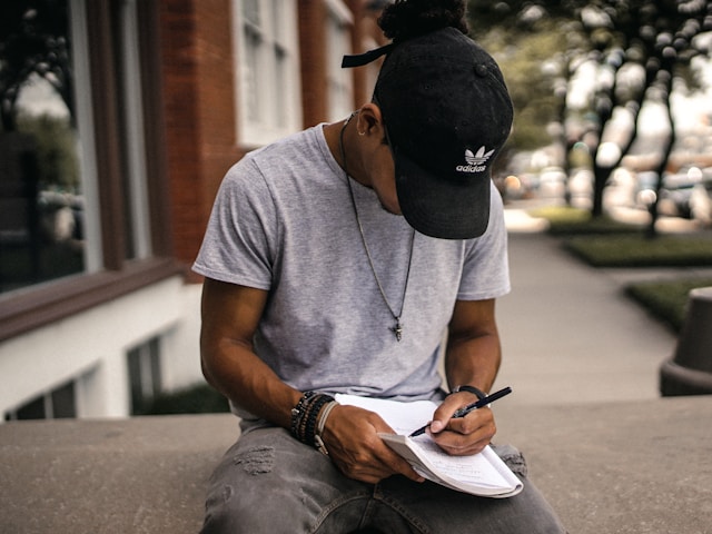 A person with a gray shirt and a black cap sits on a parapet wall and writes on a book. 