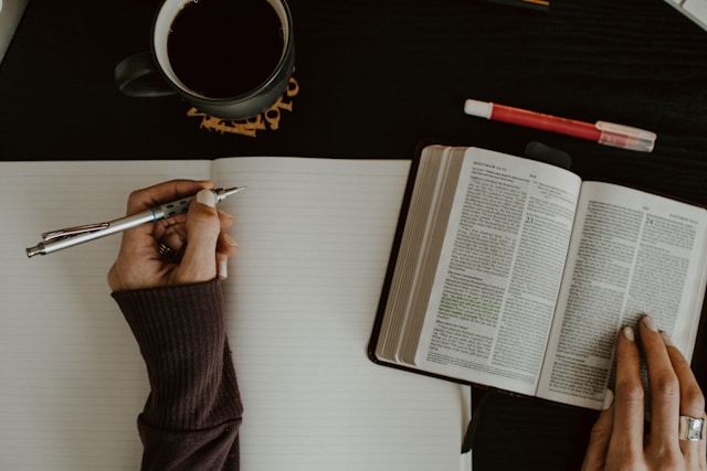 A person in a brown sweater holds an open book in one hand and a pen in the other hand. 