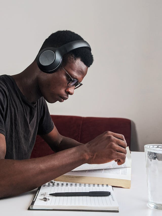 A man with a black shirt and black-colored headphones reads a book. 