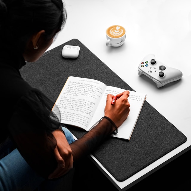 A woman writes on a book with a red-colored pen on a white table with a gray cloth.