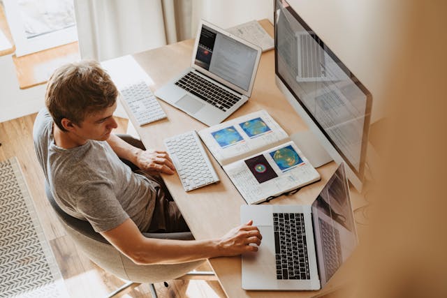 Aerial photo of a man sitting at a desk using multiple computers with notes in front of him. 