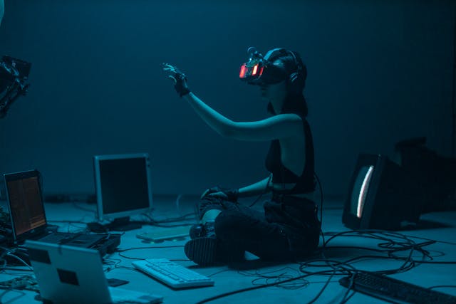 A girl is sitting in a dark room with computers around her, wearing a VR headset. 