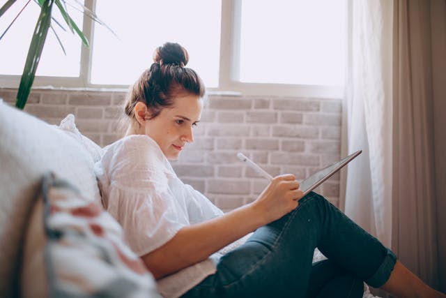 A woman sitting on a couch with her iPad, making edits on an AI-written essay. 