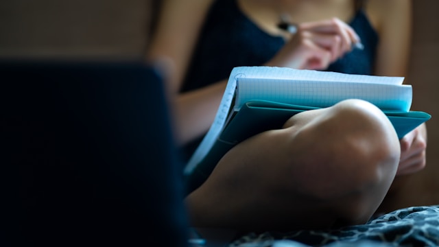 A woman sits on a bed and writes on a sheet of paper. 