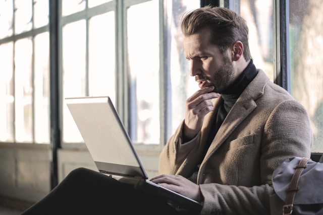 A person with a brown sweater holds a gray laptop and looks puzzled. 