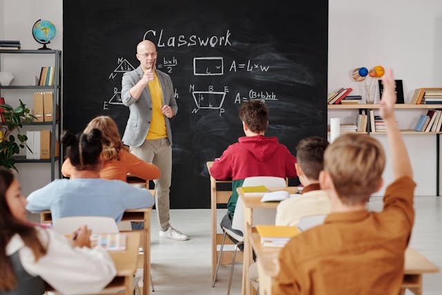 A teacher with a gray suit asks a question, and a student with an orange shirt raises their right hand. 