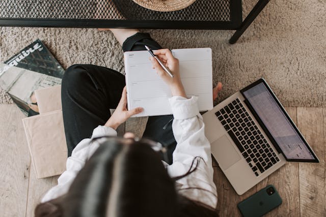 An overhead shot of a woman taking notes and working on her laptop on a couch. 