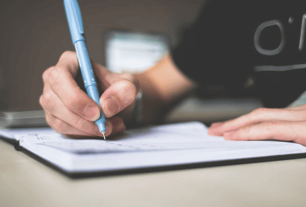 Close up of a hand holding a blue pen, grading an essay by hand in a notebook.