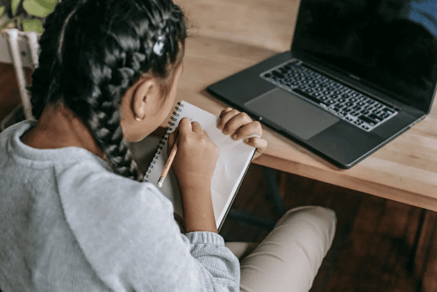 Over the shoulder view of a girl with braids writing in a notebook and looking at a laptop screen.