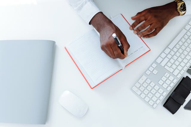 An aerial shot of a person’s hands writing in a notebook next to a computer. 