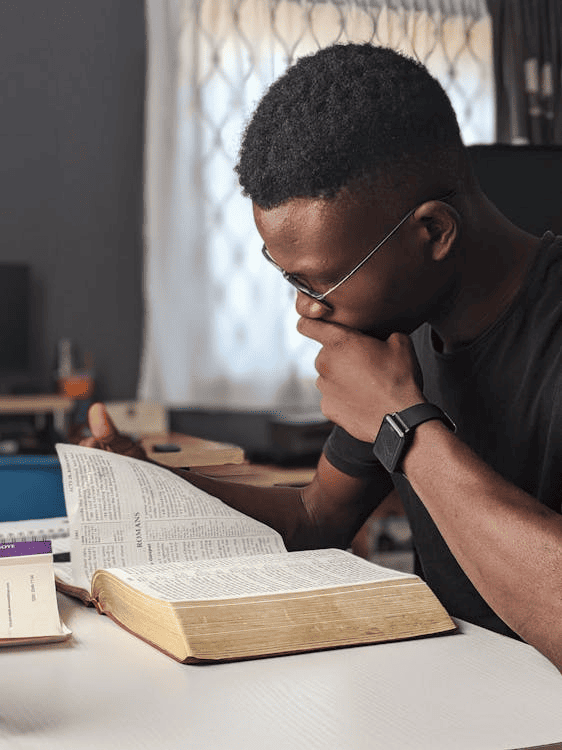 A person writing with a blue pen in a notebook with a laptop, a cup of coffee, and some books next to them.