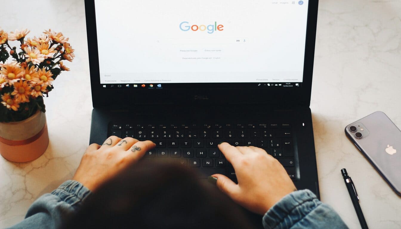 A person using Google on a laptop that is next to a plant and a smartphone.