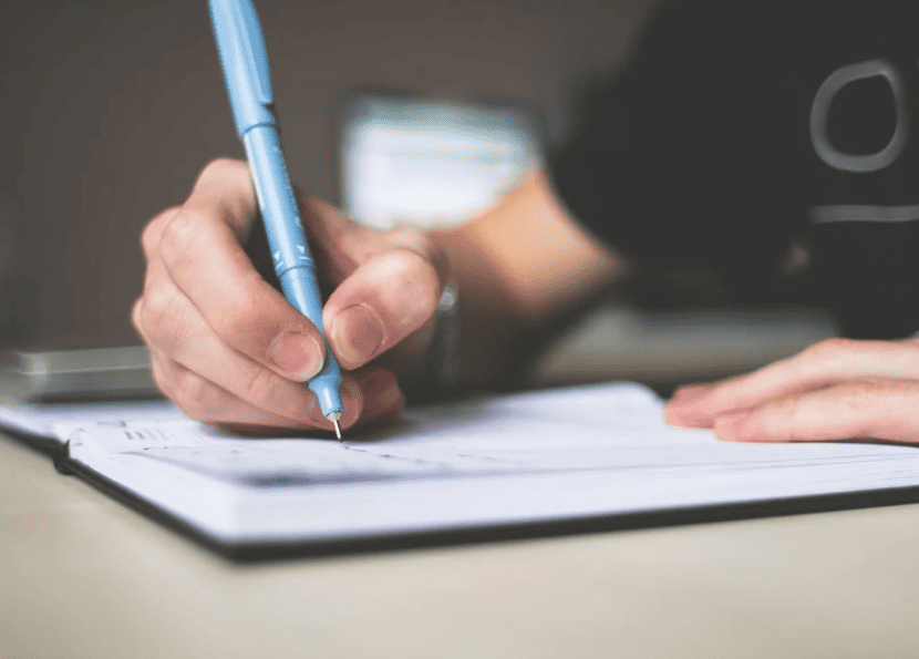 A person writing in a notebook with a blue pen. 