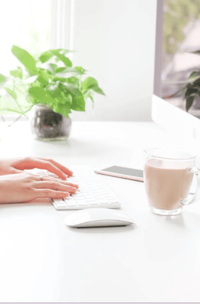 A person sitting at a desk with a plant and a cup of coffee next to them and typing on a keyboard. 