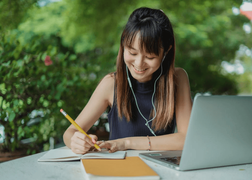 A student smiling while writing with a yellow pencil with a laptop nearby. 