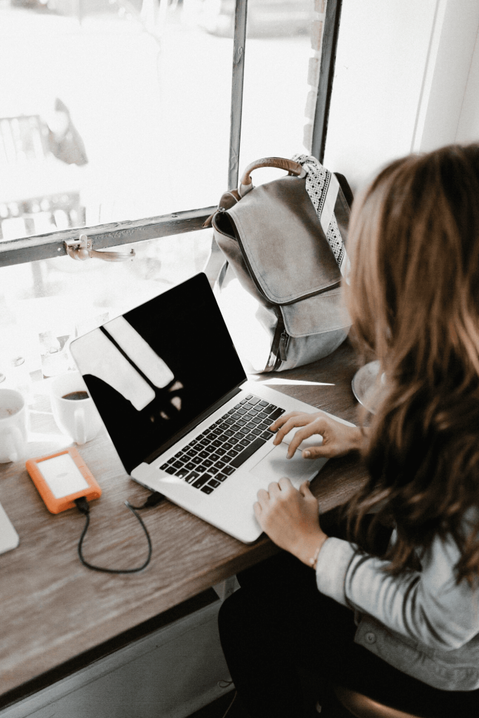 A woman typing on a laptop while sitting on a wooden desk near a window.