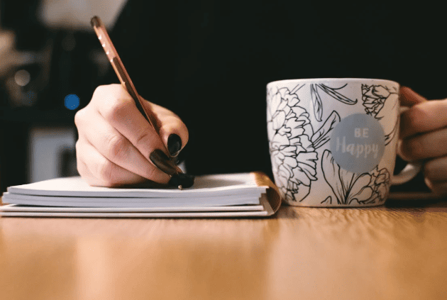 A person writing in a notebook while holding a cup. 