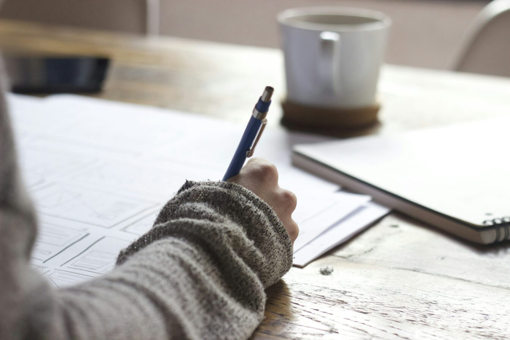 A person at a wooden desk writing with a pen on a piece of paper. 