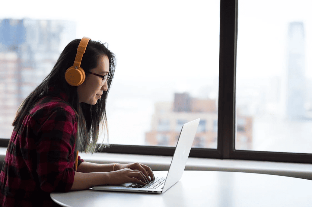 A woman with orange headphones typing on a laptop while sitting near a big window.