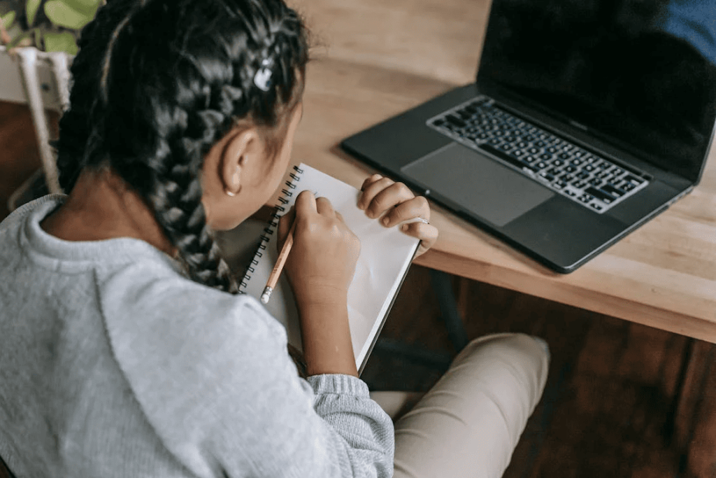 A girl sitting on a desk and writing in a notepad with a laptop in front of her.