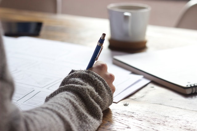 A person writing an essay with pen and paper at a wooden desk. 