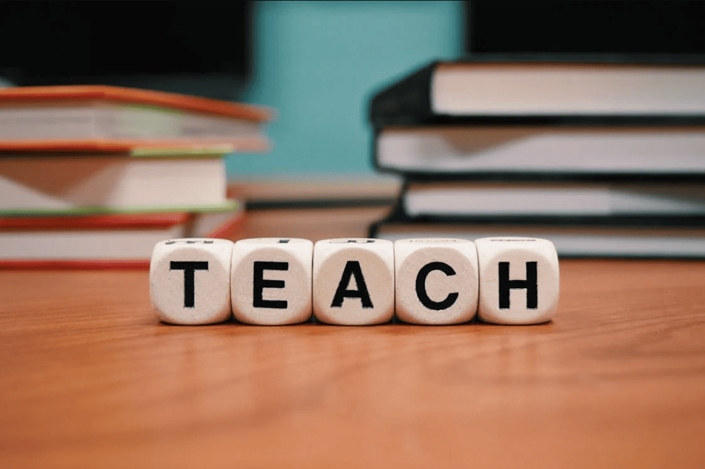 The word "Teach" made out of cubes on a desk with some books in the background.