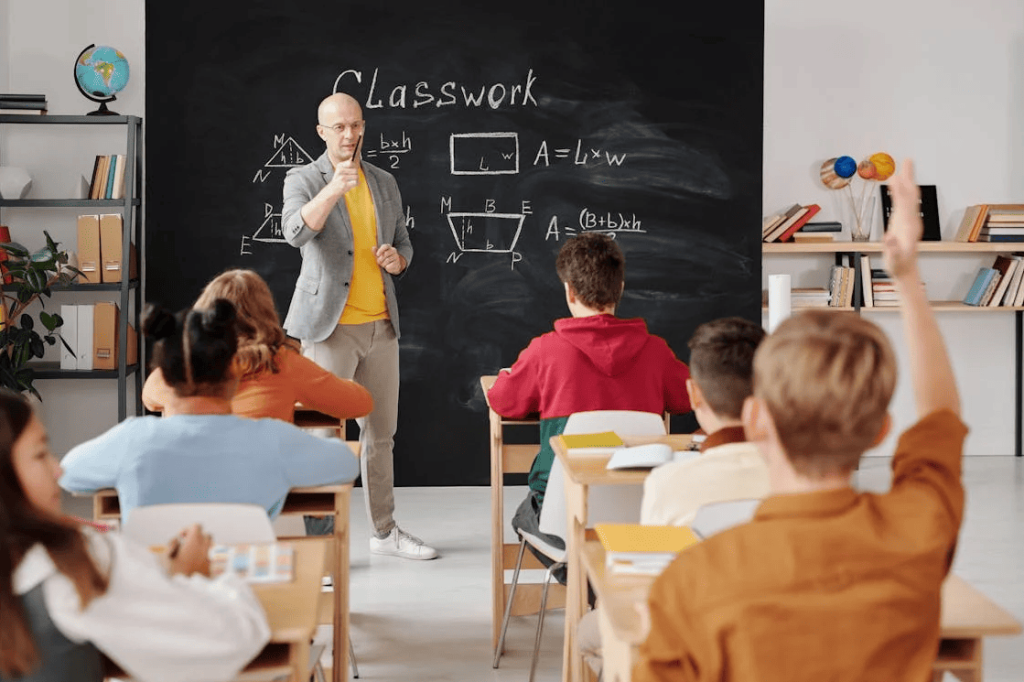 A teacher in front of a blackboard and students sitting on their desks with some raising their hand.