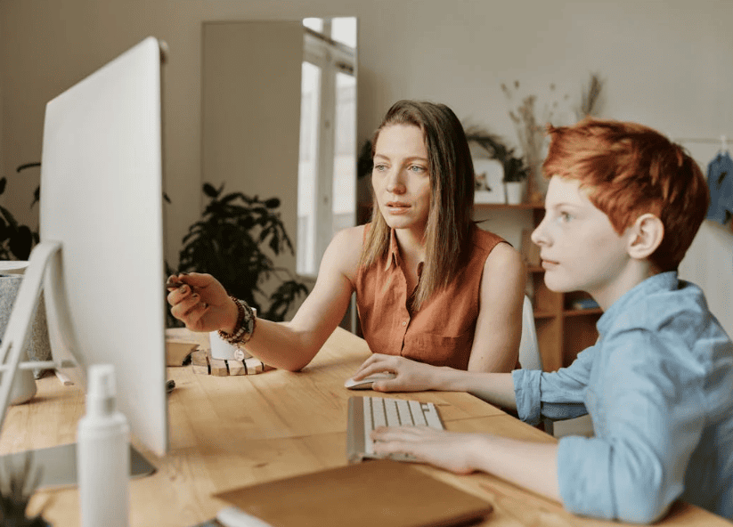 A woman and a young boy focus on a computer screen, working together at a wooden desk in a cozy, well-lit room.