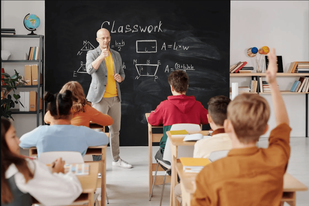 A teacher standing in front of a blackboard and pointing to a student that has their hand raised. 