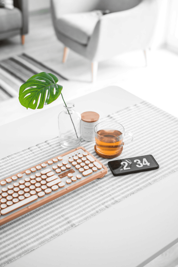 A pink computer keyboard beside a glass mug, a digital clock, and a jar with a leaf in it.