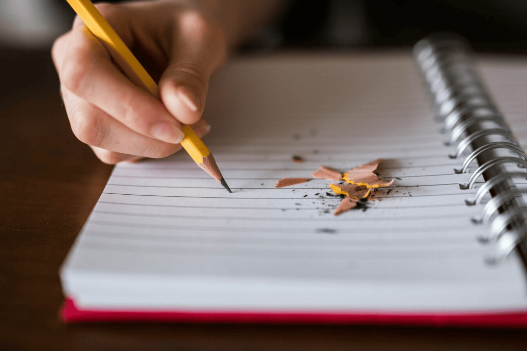 A person holding a pencil writing on top of a notebook with some pencil shavings next to it.