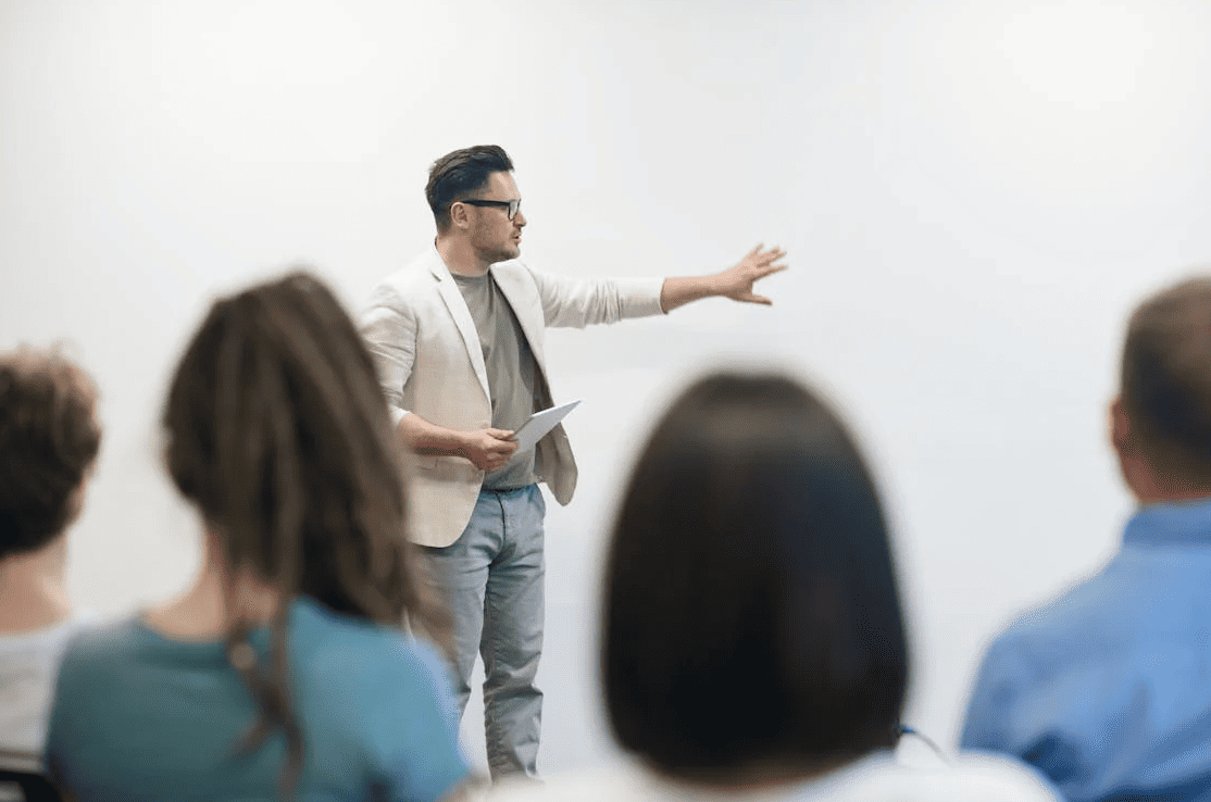 A teacher in a classroom full of students pointing to something on the board.